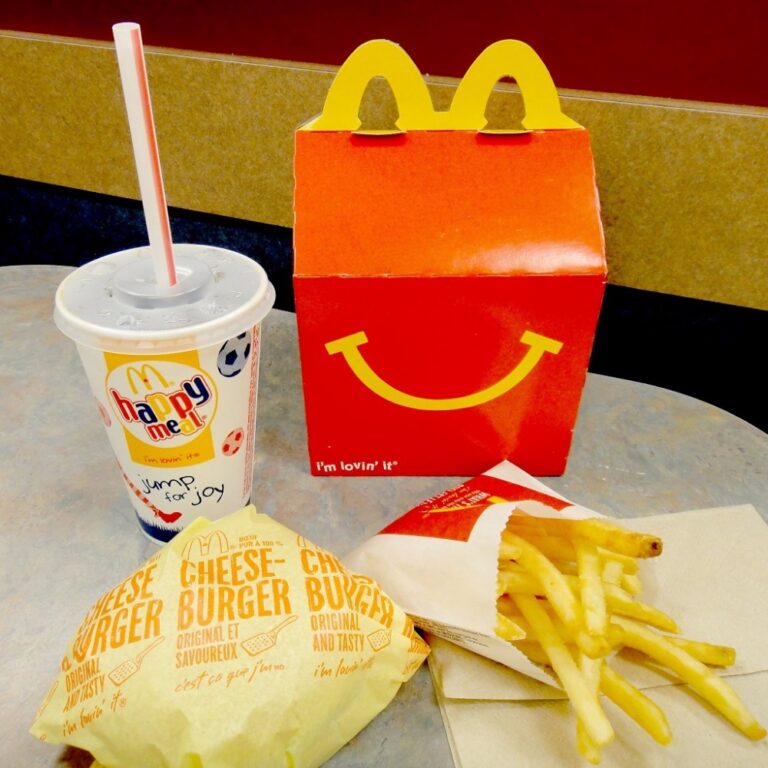 A classic McDonald’s Happy Meal displayed on a table, featuring the iconic red and yellow Happy Meal box with a smiling face design. In front of the box are a small fries in branded packaging, a wrapped cheeseburger, and a kid-sized drink cup with a straw and colorful Happy Meal graphics. The scene captures the playful and nostalgic appeal of a McDonald's meal designed for children.