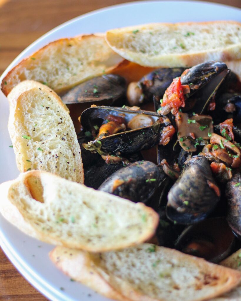 A dish of steamed mussels garnished with diced tomatoes and herbs, surrounded by slices of toasted bread, served on a white plate from Sugar Magnolia.