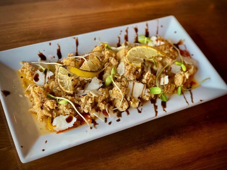 A rectangular white plate of fried squid from Sugar Magnolia Bistro & Bar, garnished with thin lemon slices, bean sprouts, shaved Parmesan, and a drizzle of balsamic glaze. The dish is artfully arranged on a wooden table.