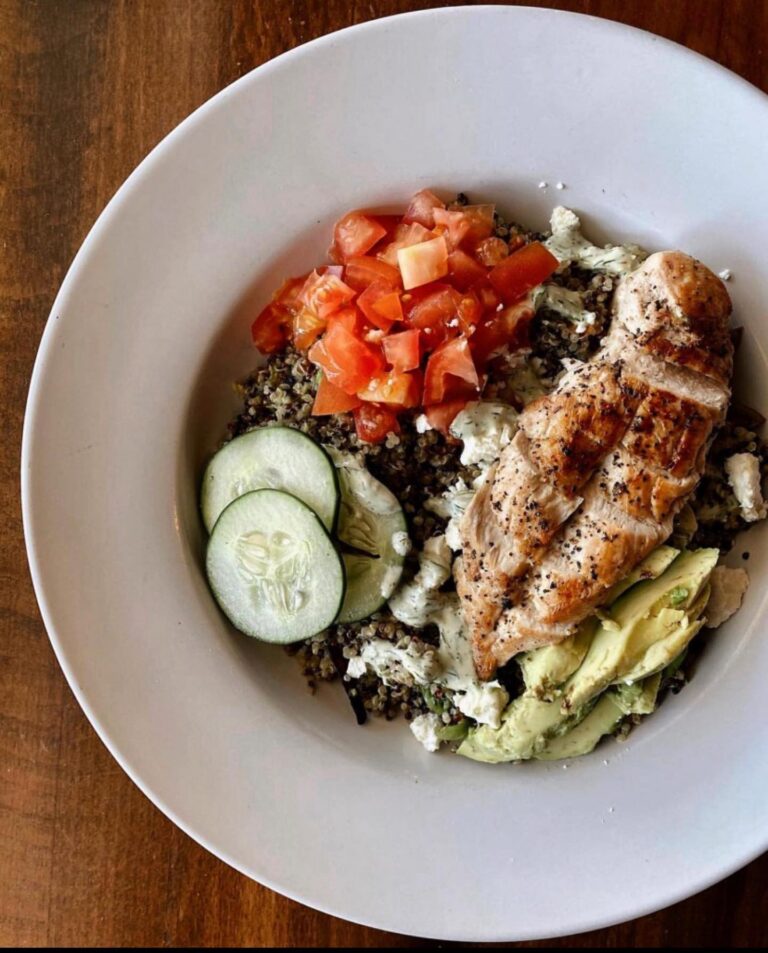 A hearty bowl from Sugar Magnolia Bistro & Bar featuring a grilled chicken breast served over a bed of seasoned rice and quinoa, topped with chopped tomatoes, cucumber slices, and avocado. The dish is presented in a wide white bowl on a wooden table.