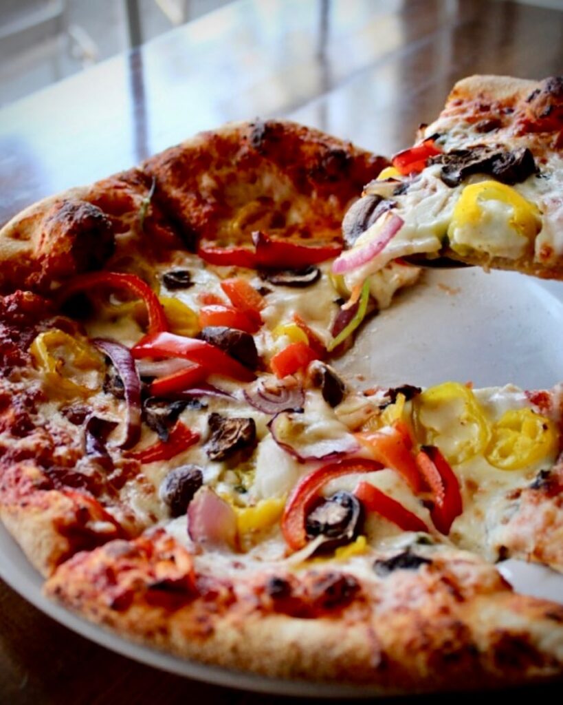 A vibrant vegetable pizza from Sugar Magnolia Bistro & Bar, served on a white plate. The pizza is topped with red onions, mushrooms, red and yellow bell peppers, and melted cheese, with one slice being lifted to reveal a golden, crispy crust. The background shows a softly lit wooden table.