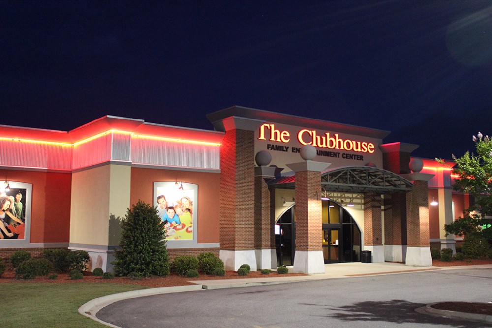 A nighttime view of "The Clubhouse," a family entertainment center. The building has bright red neon lights outlining its roof and a prominent sign with the name "The Clubhouse" in glowing letters. The entrance features brick columns and an arched canopy, with landscaped shrubs and a paved parking area in the foreground.