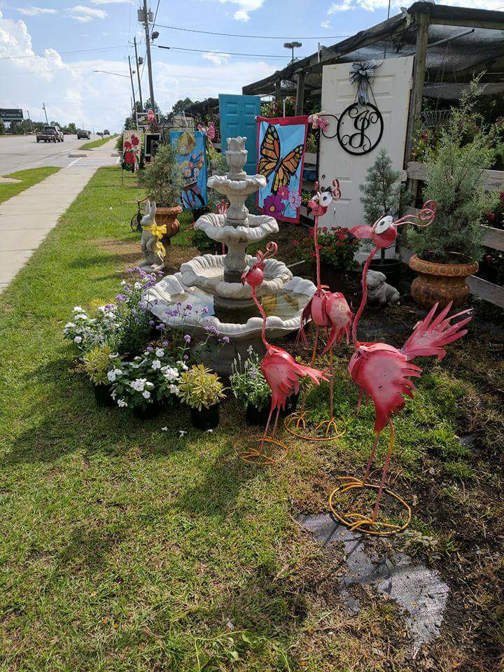 A colorful roadside garden display featuring a tiered stone fountain surrounded by vibrant potted flowers and whimsical metal flamingo sculptures. Behind the fountain are hand-painted decorative panels with bright butterflies and floral themes, along with repurposed doors and wall art. The eclectic mix of garden decor sits next to a sidewalk and busy street, creating an eye-catching and cheerful scene that invites passersby to stop and explore.