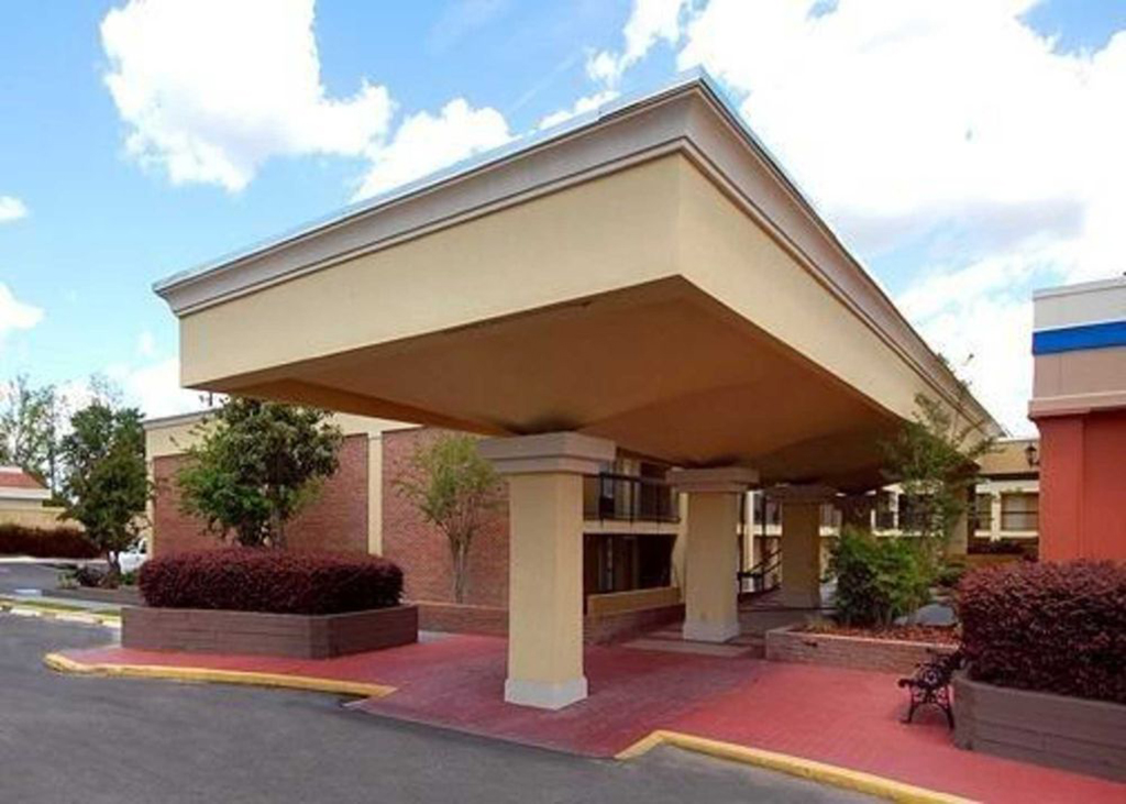 Exterior view of a hotel entrance with a large covered driveway supported by columns. The building features a combination of beige, yellow, and brick-red tones, with landscaped planters and a bench near the entrance. The sky is partly cloudy, creating a bright and welcoming atmosphere.