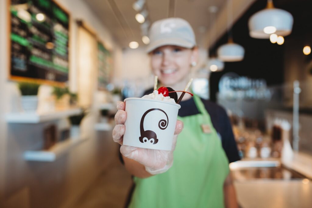 A Rolling Monkey employee wearing a gray cap and green apron smiles while holding out a cup of rolled ice cream topped with whipped cream, a cherry, and wafer sticks. The cup features the Rolling Monkey logo with a stylized monkey. The background shows a bright, modern ice cream shop interior.