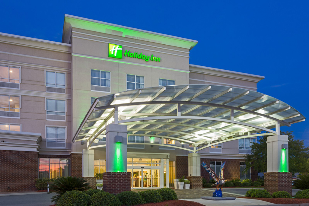 Exterior view of the Holiday Inn hotel at dusk, featuring a modern glass-covered entrance with green accent lighting. The four-story building has beige and brick walls, large windows, and a well-lit lobby visible through the glass doors. A colorful eagle sculpture is displayed near the entrance, surrounded by landscaped greenery.
