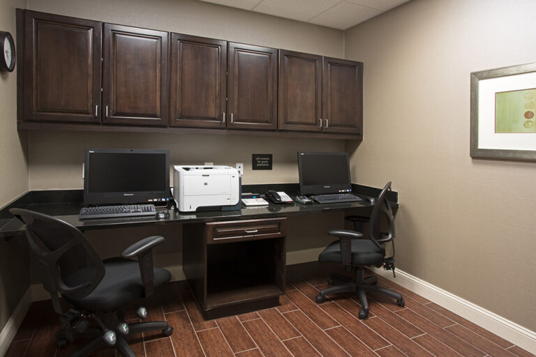A small business center with two black office chairs, two desktop computers, a white printer, and a phone on a black countertop. The room features dark wooden cabinets above the workspace, wood-patterned tile flooring, beige walls, and a framed abstract artwork on the right wall.