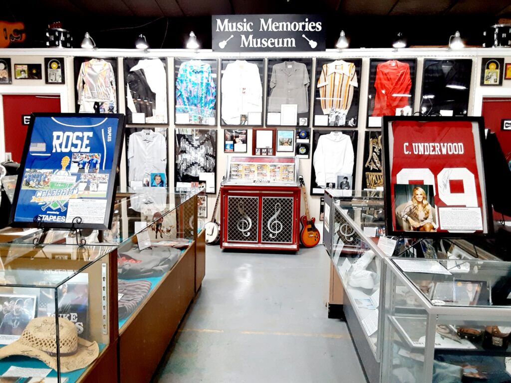 Interior view of the Music Memories Museum showcasing framed stage outfits worn by musicians, a central vintage jukebox, and a guitar on display. Two signed jerseys—one blue labeled “ROSE” and one red labeled “C. UNDERWOOD” with a photo of Carrie Underwood—are prominently featured. Glass display cases on either side hold memorabilia, including hats, photographs, and clothing, under bright overhead lighting.