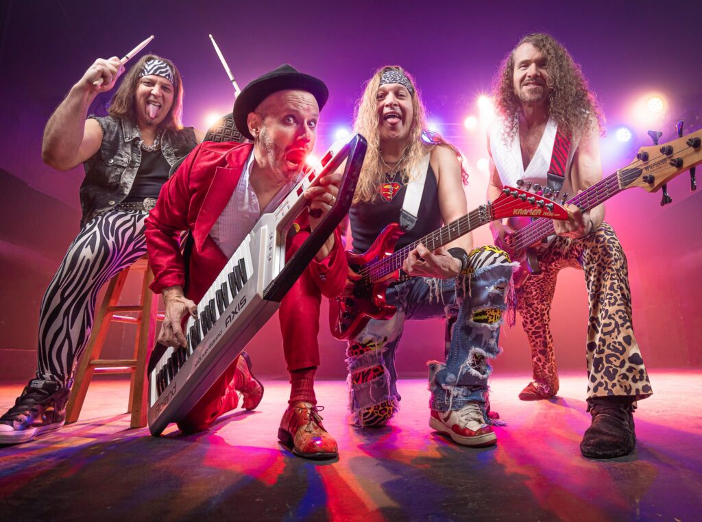 A high-energy promotional photo of The Breakfast Club band on stage, dressed in flashy, 1980s-inspired outfits. One member is kneeling with a keytar, another holds a red electric guitar, while the other two play bass and drumsticks. They wear animal print pants, ripped jeans, and headbands, surrounded by bright stage lights and a vibrant performance atmosphere.