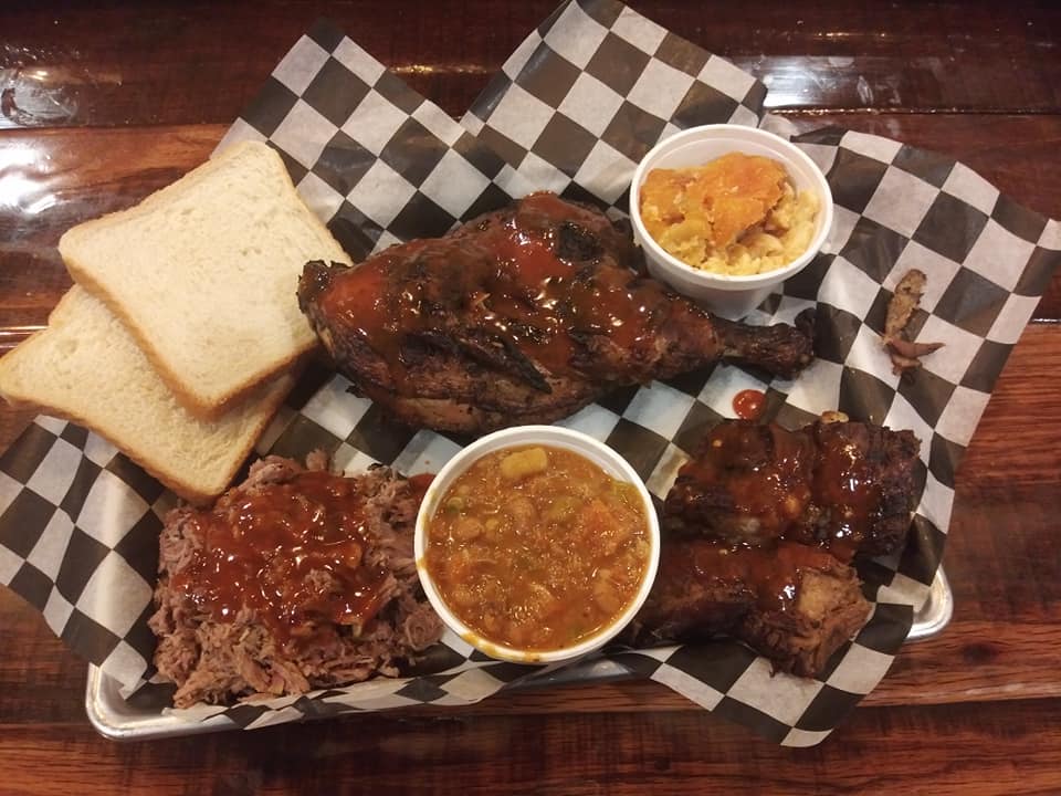 A platter from Baby CJ's Rib Shack featuring barbecue chicken, ribs, pulled pork topped with sauce, slices of white bread, and sides of baked beans and peach cobbler, served on checkered paper.