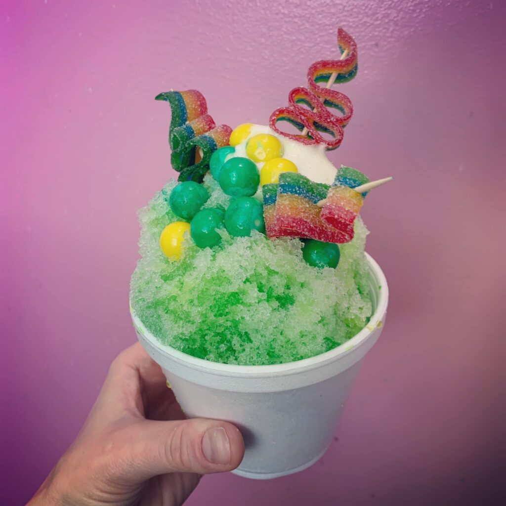 A vibrant green snow cone from Boro Sno, served in a white styrofoam cup and topped with green apple syrup. The treat is creatively decorated with colorful candy, including rainbow sour strips, gumballs in yellow and green, and twisted gummy ribbons skewered on sticks. The background features a pink-purple gradient wall, and a hand is holding the cup from the bottom.