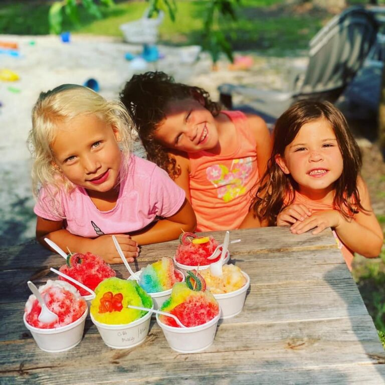 Three young girls smile at a picnic table outdoors on a sunny day, each wearing colorful summer clothes. In front of them are several colorful snow cones topped with various candies, served in white cups with plastic spoons. The background features a sandbox and green grass.