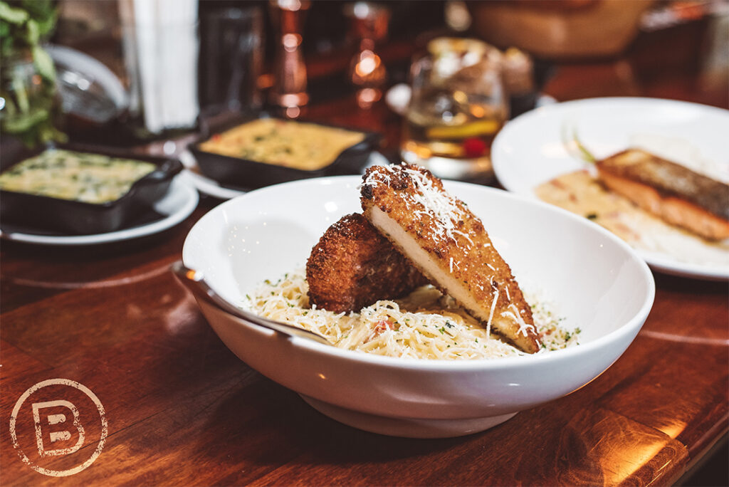 A white bowl filled with creamy noodles topped with two crispy, golden-brown fried chicken breasts, garnished with shredded cheese and herbs. The dish is set on a polished wooden table at Bull Barrel Steakhouse, surrounded by other plated meals and drinks in a cozy, upscale dining setting.