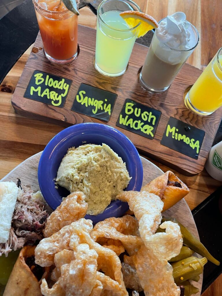 A colorful display of food and drinks at Dolan's BBQ. At the top, a wooden flight board holds four drinks labeled "Bloody Mary," "Sangria," "Bush Wacker," and "Mimosa," each in a small glass with garnish. Below, a wooden platter features a variety of snacks including pork rinds, spring rolls, pickles, pulled pork, and a scoop of dip in a blue bowl.