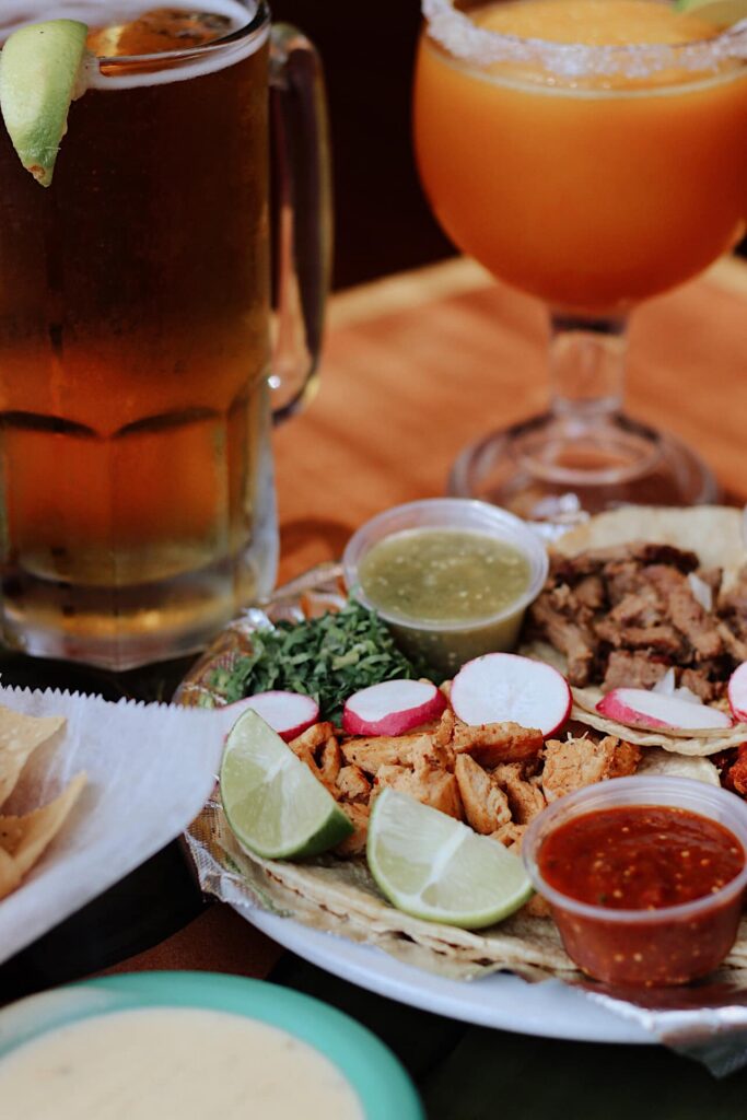 A vibrant promotional photo of El Jalapeno’s offerings featuring a plate of fresh tacos garnished with lime wedges, radish slices, cilantro, and two types of salsa. The background includes a frosty mug of beer and a frozen margarita, both garnished with lime. A basket of tortilla chips and a bowl of queso are partially visible, suggesting a full and festive Mexican dining experience.