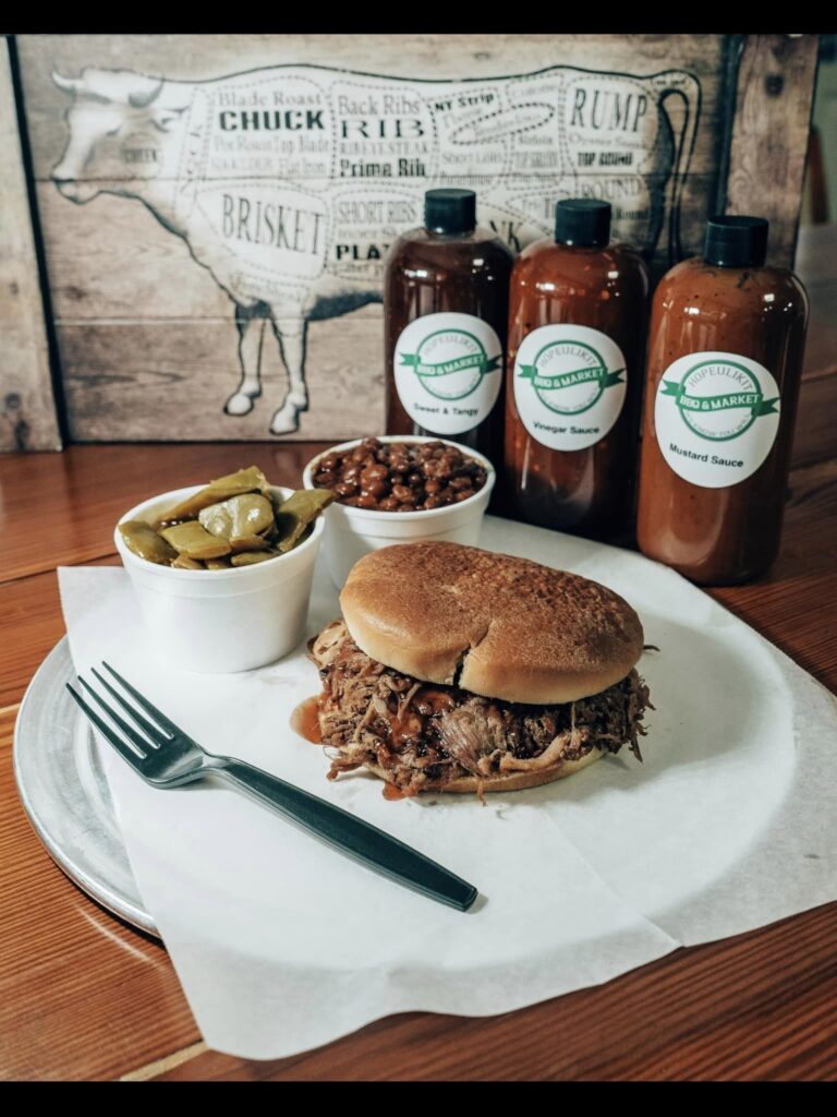 A promotional image for Hopeulikit BBQ & Market featuring a pulled pork sandwich on a bun, served with a side of baked beans and pickles in white cups. In the background are three bottles of Hopeulikit BBQ sauces labeled "Sweet & Tangy," "Vinegar Sauce," and "Mustard Sauce." A butcher diagram of a cow is displayed on a wooden board in the background, reinforcing the BBQ theme.