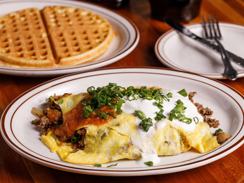 A hearty breakfast plate from Loc's Chicken & Waffles featuring a loaded omelet filled with seasoned ground beef, sautéed onions, and melted cheese. The omelet is topped with a generous serving of sour cream, salsa, and freshly chopped green onions. In the background, a golden brown waffle, cut into four sections, sits on a separate plate. A small plate with a fork and knife rests nearby on a wooden table, completing the classic diner-style breakfast presentation.