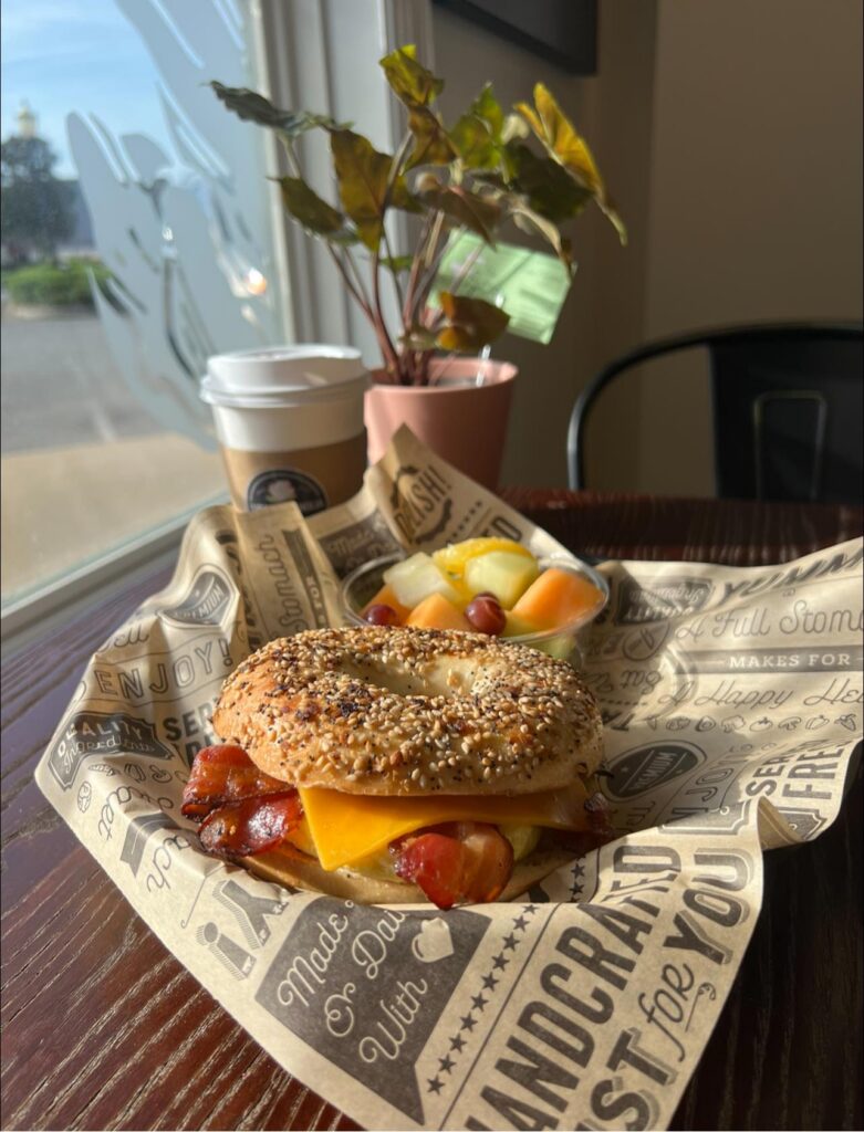 A breakfast bagel sandwich from Sugar Magnolia Bistro & Bar, featuring crispy bacon, melted cheddar cheese, and a fluffy egg between a toasted everything bagel. Served on branded deli paper alongside a cup of coffee and a side of fresh fruit, all set on a wooden table near a sunny window with a potted plant in the background.