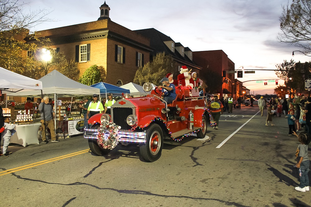Santa Claus waves from the back of a vintage red fire truck decorated with Christmas lights and a wreath during the Statesboro holiday parade. The event takes place at dusk in downtown Statesboro, with people lining the street and vendors set up under white tents. The festive atmosphere includes holiday lights, costumed characters, and children watching the parade.