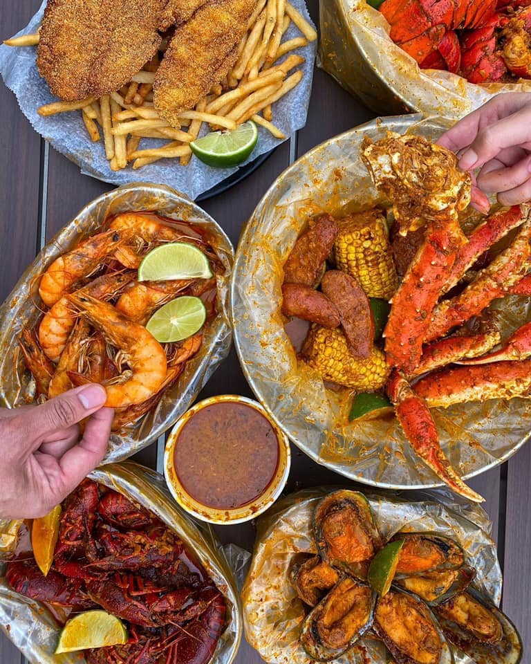 A vibrant seafood spread from Red Claw Juicy Seafood & Bar. The image shows various seafood dishes including boiled shrimp, crawfish, mussels, crab legs with corn and sausage, and fried catfish with fries. Each dish is served in clear plastic bags coated with rich, seasoned sauce. A cup of dipping sauce sits in the center, and lime wedges are used as garnish throughout the spread. Hands are seen reaching in to enjoy the food, emphasizing the communal, hands-on dining experience.
