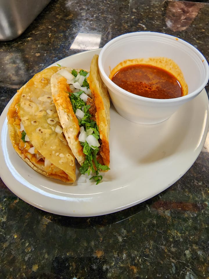 A plate of La Casita's Carne Asada Tacos served with a side of rich, red dipping broth. The two tacos are filled with juicy carne asada, chopped onions, and fresh cilantro, all tucked into golden, crispy tortillas. The broth, likely a savory consommé, is presented in a white cup, perfect for dipping the flavorful tacos. The dish is placed on a white plate over a dark, glossy countertop.