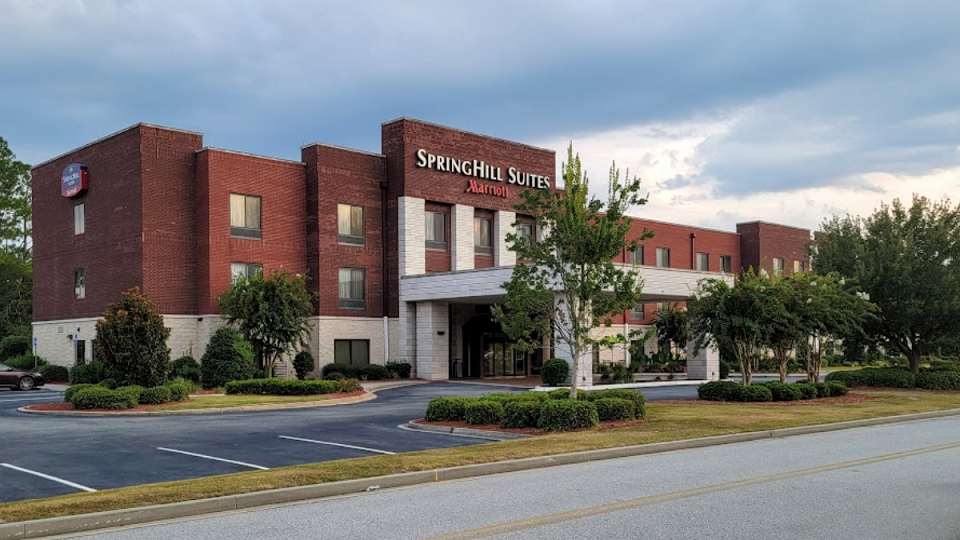 Exterior photo of the SpringHill Suites by Marriott in Statesboro, showcasing a modern three-story brick building with a covered entrance driveway, manicured landscaping, and clearly marked parking spaces. The hotel's signage is prominently displayed at the top of the building. A partly cloudy sky adds a calm, inviting ambiance to the scene.