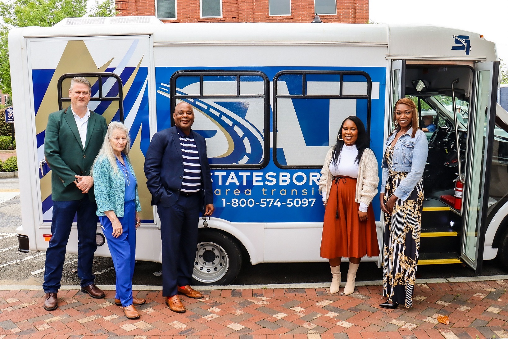 Start-up photo of Statesboro's Public Transit System featuring five individuals standing in front of a branded Statesboro Area Transit (SAT) shuttle bus. The group is dressed in professional and stylish attire, smiling for the photo on a brick-paved sidewalk. The bus displays the SAT logo and contact number, with a building visible in the background, signifying a celebratory or launch event.