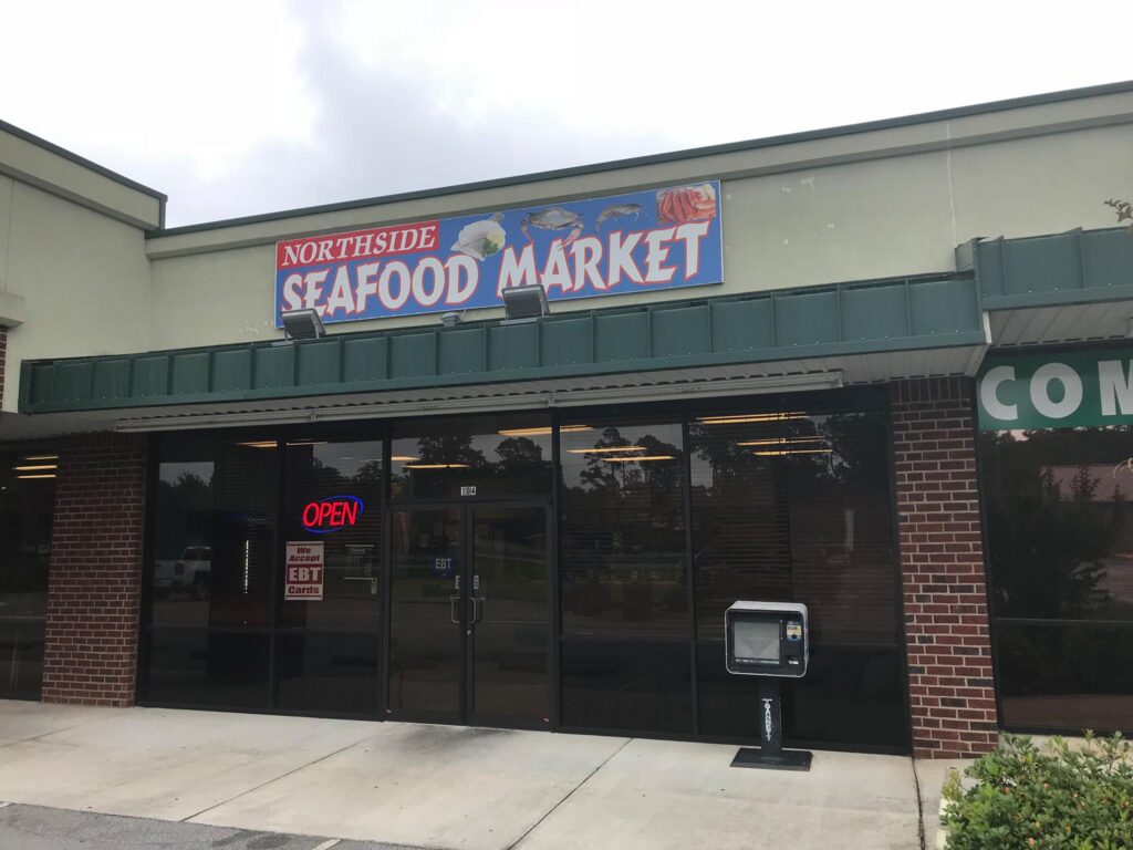 The exterior of Northside Seafood Market, a seafood store located in a strip mall. The storefront features a large sign with the name 'Northside Seafood Market' in bold red and blue lettering, along with images of seafood such as fish, shrimp, and crabs. The entrance has glass doors with a neon 'OPEN' sign and a notice indicating that EBT cards are accepted. A small newspaper vending machine is positioned outside. The building has a brick and green awning design, and the reflection in the glass windows shows a parking lot with trees in the background.