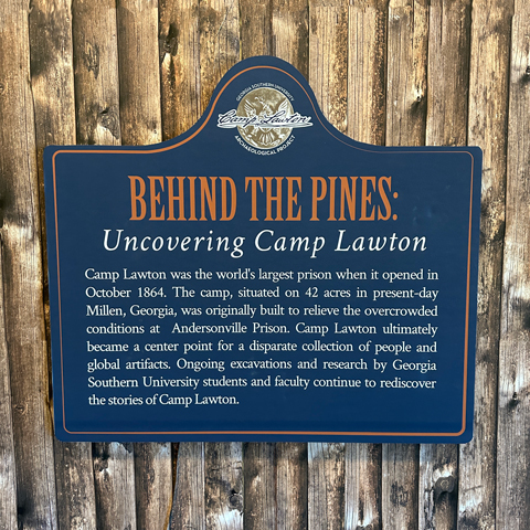 A historical sign titled 'BEHIND THE PINES: Uncovering Camp Lawton,' mounted on a wooden fence. The sign provides information about Camp Lawton, stating that it was the world's largest prison when it opened in October 1864. Located on 42 acres in present-day Millen, Georgia, the camp was originally built to relieve overcrowding at Andersonville Prison. It later became a significant site for people and artifacts. The sign also highlights the ongoing excavations and research conducted by Georgia Southern University students and faculty to continue uncovering the stories of Camp Lawton. The sign has a navy blue background with gold and white text, topped with an emblem that features the name 'Camp Lawton' and an image of pine trees.