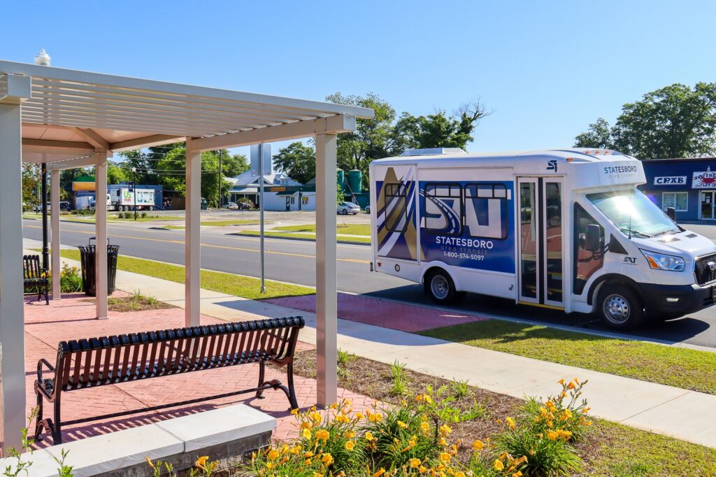 A visual of the public transit system in Statesboro, featuring a white and blue Statesboro Area Transit (SAT) shuttle bus parked at a designated bus stop. The stop includes a covered seating area with benches and a sidewalk bordered by blooming yellow flowers. Businesses and residential buildings are visible across the street under a clear blue sky.