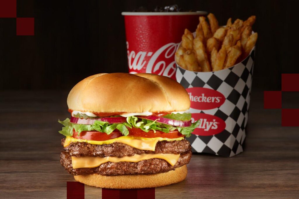 A classic fast-food meal from Checkers featuring a double cheeseburger, seasoned fries, and a Coca-Cola drink. The burger is stacked with two beef patties, melted American cheese, crisp lettuce, fresh tomato slices, red onion rings, pickles, and ketchup, all inside a soft toasted bun. In the background, a container of Checkers’ signature seasoned fries is placed in a black-and-white checkered cup with the Checkers and Rally’s logo, alongside a red Coca-Cola cup filled with ice-cold soda. The dark wooden surface and subtle red square accents in the background enhance the bold and appetizing presentation.