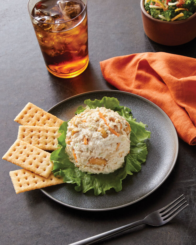 A serving of Chicken Salad Chick's Jalapeño chicken salad, plated on a dark dish with a fresh lettuce leaf. The creamy chicken salad contains shredded chicken, jalapeños, and other seasonings, creating a flavorful dish. It is accompanied by a side of crispy saltine crackers. In the background, a glass of iced tea with condensation sits on a dark stone surface, next to a small bowl of broccoli salad. An orange napkin adds a warm, inviting touch to the presentation, and a silver fork rests nearby, ready for dining.