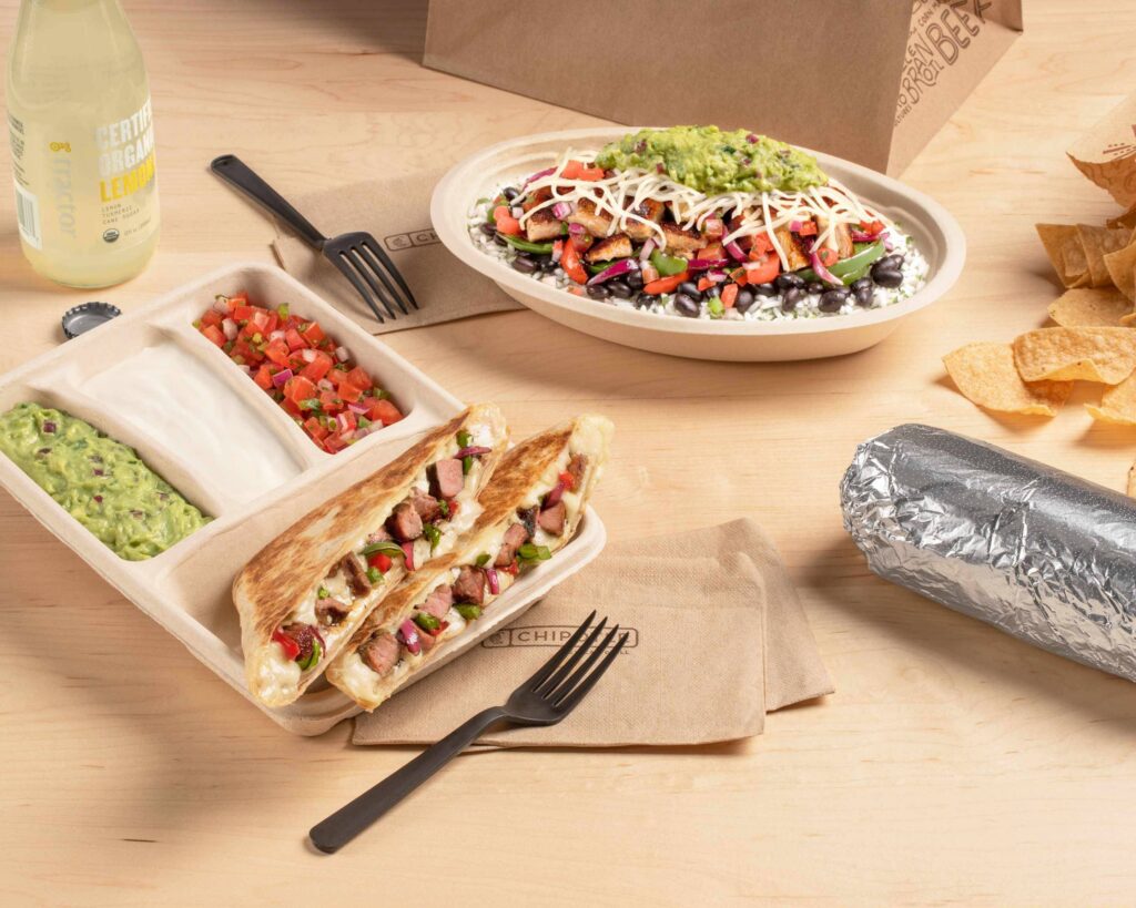 A spread of Chipotle's various food options arranged on a light wooden table. The meal includes a quesadilla filled with grilled steak, cheese, and fresh vegetables, served in a divided tray with sides of guacamole, sour cream, and pico de gallo. In the background, there is a burrito bowl featuring black beans, white rice, grilled vegetables, pico de gallo, shredded cheese, and a generous scoop of guacamole. To the side, a foil-wrapped burrito sits next to a portion of crispy tortilla chips. A bottle of organic lemonade is also included, along with Chipotle-branded napkins and utensils, completing the fast-casual dining experience.