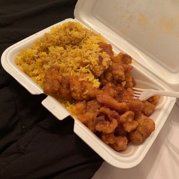 A takeout container from Great Wall featuring a serving of orange chicken and yellow fried rice. The orange chicken consists of crispy battered pieces coated in a glossy, tangy-sweet sauce. The fried rice is golden in color and appears well-seasoned. A white plastic fork rests in the container, which is placed on a soft fabric surface, suggesting a casual dining setting.
