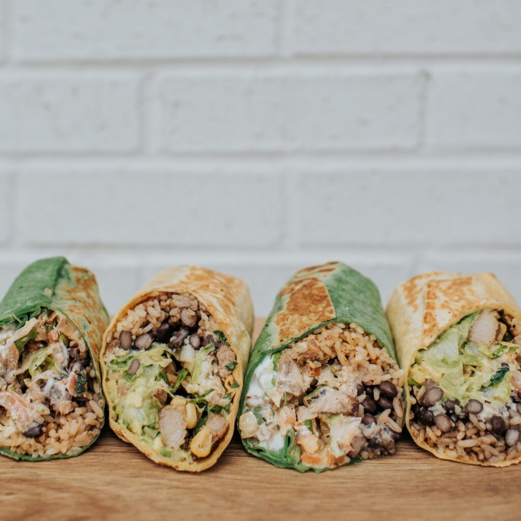 A close-up of four freshly made burritos from Surcheros Fresh Grill, cut in half and arranged on a wooden surface. Each burrito is packed with seasoned rice, black beans, grilled chicken, lettuce, guacamole, and other fresh ingredients. Two of the burritos are wrapped in spinach tortillas, while the other two are in traditional flour tortillas, all toasted to a golden brown. The background features a white brick wall, highlighting the vibrant colors and textures of the burritos.