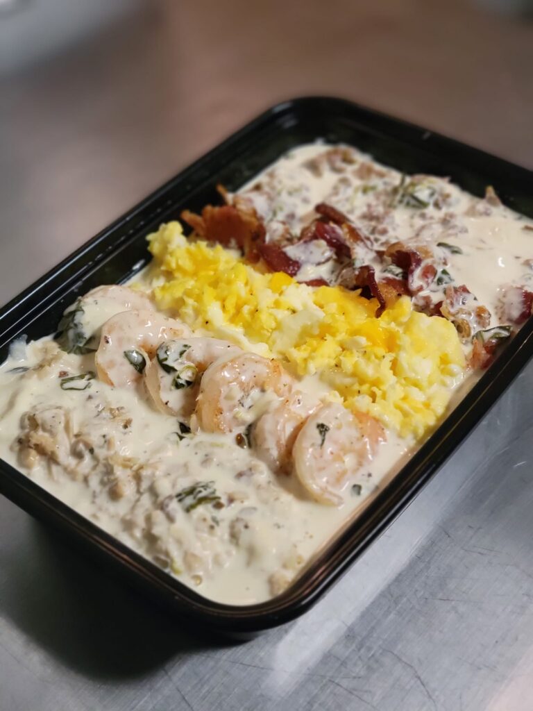 A takeout container from Tasty Crave filled with a rich and creamy dish featuring shrimp, scrambled eggs, crispy bacon, and a savory sauce. The dish is divided into sections, with plump shrimp covered in a creamy white sauce with herbs, a portion of fluffy yellow scrambled eggs, and crispy bacon pieces over what appears to be a bed of grits or hash browns, all generously topped with sauce. The black container is placed on a stainless steel surface, emphasizing the hearty and indulgent meal.