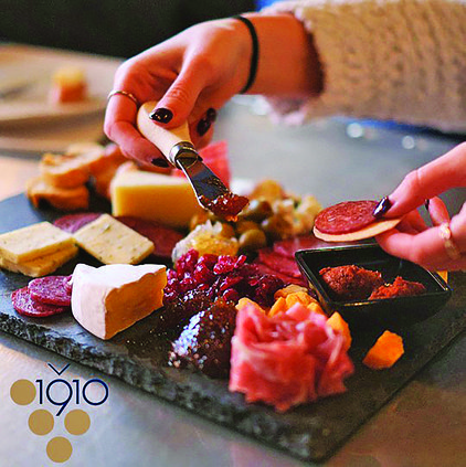 A beautifully arranged charcuterie board at Vino 1910, featuring an assortment of fine cheeses, cured meats, olives, fruit preserves, and artisan crackers on a dark slate serving platter. A person with manicured nails and elegant rings is seen spreading a rich preserve onto a cracker, while holding a slice of cured meat in the other hand. The warm lighting creates an inviting atmosphere, enhancing the textures and colors of the gourmet selection. The 'Vino 1910' logo with a stylized grape cluster is visible in the lower-left corner, complementing the sophisticated presentation of this wine and charcuterie experience.
