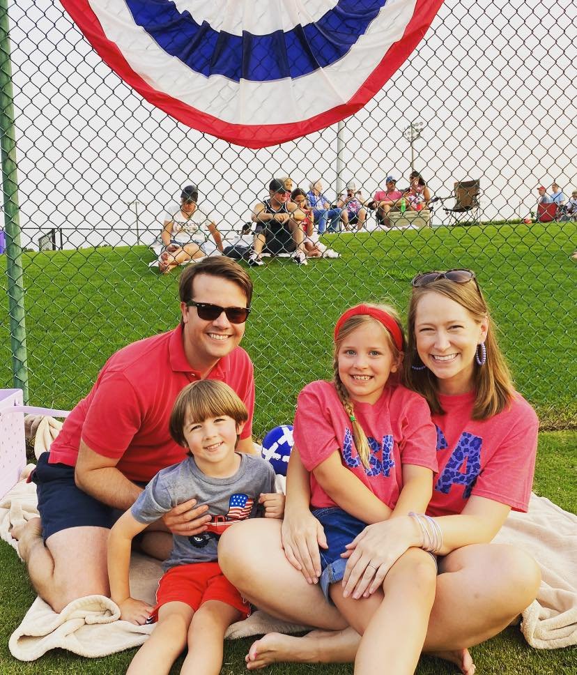 A smiling family of four poses on a blanket at the Firecracker Festival in front of a chain-link fence decorated with red, white, and blue bunting. The father wears a red polo and sunglasses, while the mother and daughter wear matching "USA" shirts. The young boy sits in front wearing a gray shirt with an American flag patch and red shorts. Behind them, festivalgoers sit on a grassy hill, enjoying the festive atmosphere