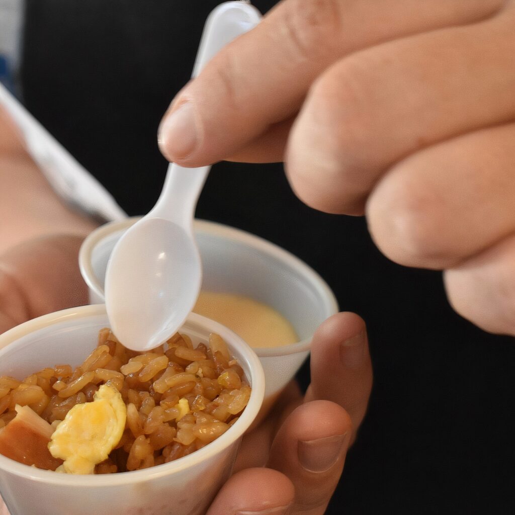 Close-up of a person holding small sample cups of food at the Wells Fargo Taste of Downtown event. One cup contains fried rice with egg and meat, and the other appears to contain a creamy soup. A plastic spoon is poised above the rice, ready to eat.