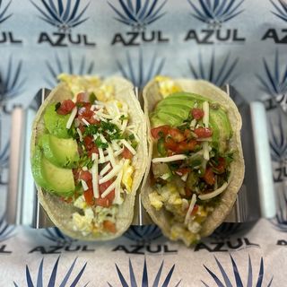Two breakfast tacos filled with scrambled eggs, sliced avocado, shredded cheese, pico de gallo, and chopped herbs, served in a metal taco holder. The background features a branded paper liner with the logo "AZUL Tex-Mex Express" and agave illustrations.