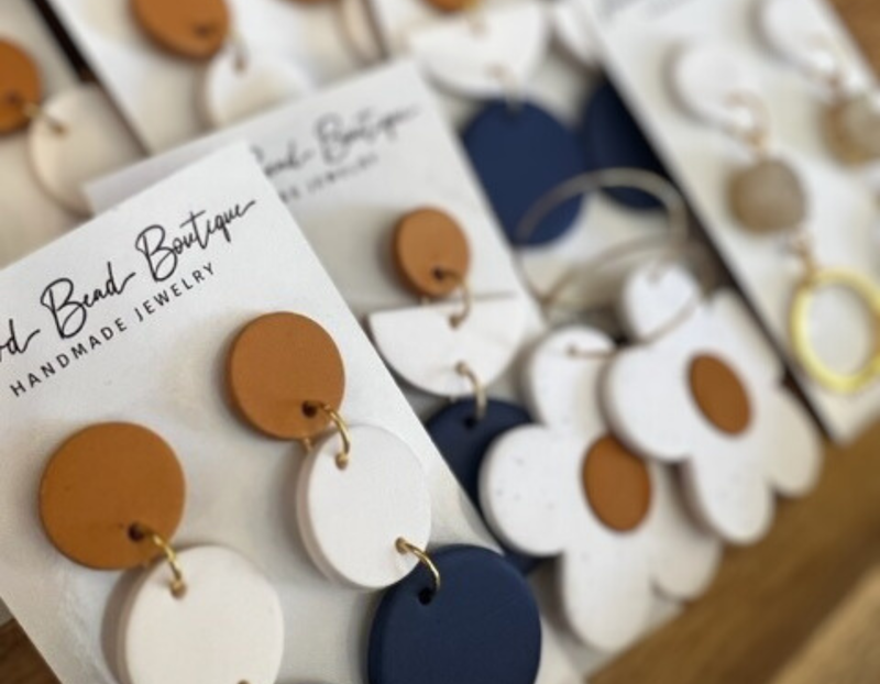 A display of handmade earrings from Wild Bead Boutique, featuring colorful geometric and floral designs. The earrings are made of clay or similar material in shades of mustard yellow, white, navy blue, and beige, arranged neatly on branded display cards.