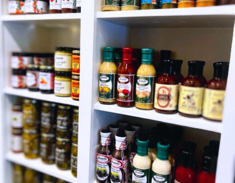 A display of neatly arranged shelves stocked with various sauces, jams, and condiments in glass and plastic bottles. The labels include fruit and vegetable illustrations, suggesting flavors like peach, apple, and garlic. The shelving unit is white, creating a clean and organized appearance.