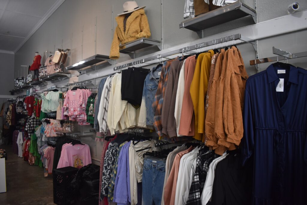 A clothing display inside Collective & Vine, showcasing a variety of stylish women’s apparel including jackets, sweaters, blouses, and dresses in various colors and fabrics. Shelves above hold purses and folded garments, while the wall-mounted racks beneath display hanging outfits in a neatly arranged boutique setting.