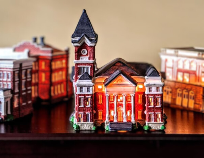 A detailed miniature model of a historic red-brick building with a tall clock tower, glowing warmly from interior lights. The structure resembles a courthouse or civic building and is surrounded by other illuminated miniature buildings, all arranged on a dark surface with a soft, neutral background.