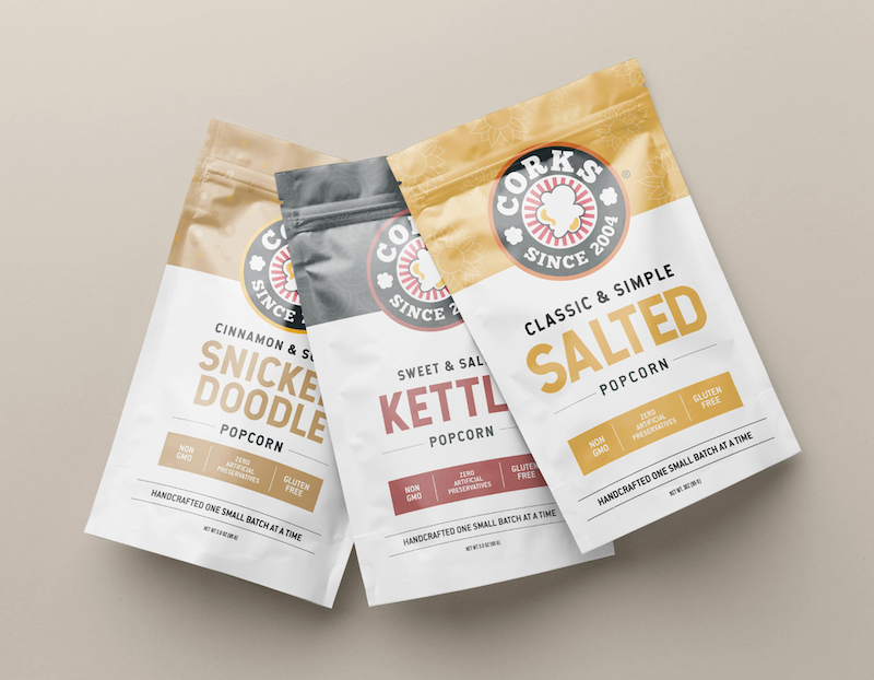 Three varieties of Corks popcorn bags are displayed side by side on a beige background. The flavors include "Cinnamon & Sugar Snickerdoodle," "Sweet & Salty Kettle," and "Classic & Simple Salted." Each bag features the Corks logo with a popcorn illustration, and highlights attributes like "Non-GMO," "Zero Artificial Preservatives," and "Gluten Free." The packaging notes that the popcorn is handcrafted one small batch at a time.