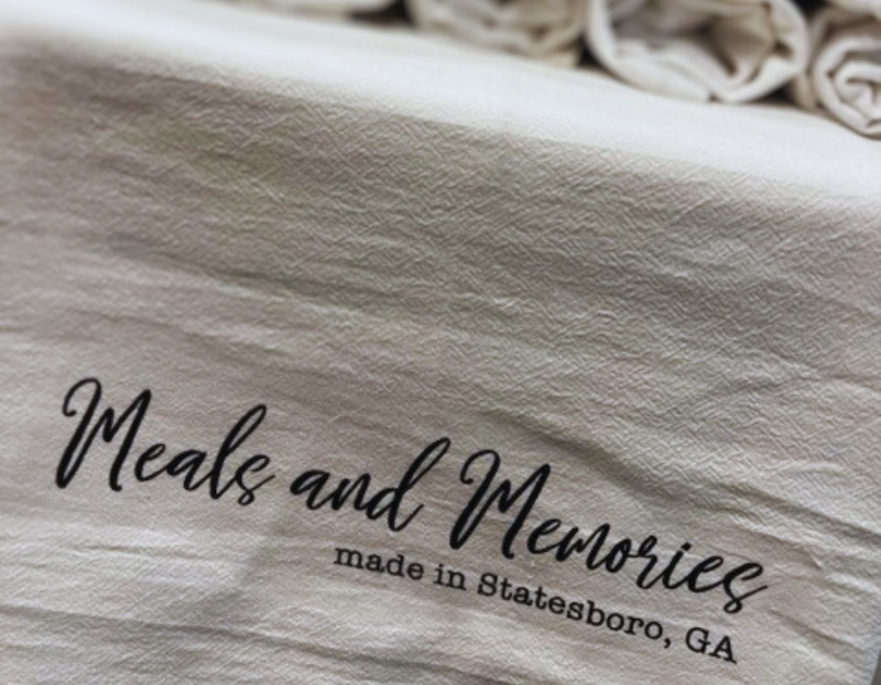 A beige cloth with the printed phrase “Meals and Memories made in Statesboro, GA” in elegant black script. Several neatly rolled matching cloths are visible in the background.