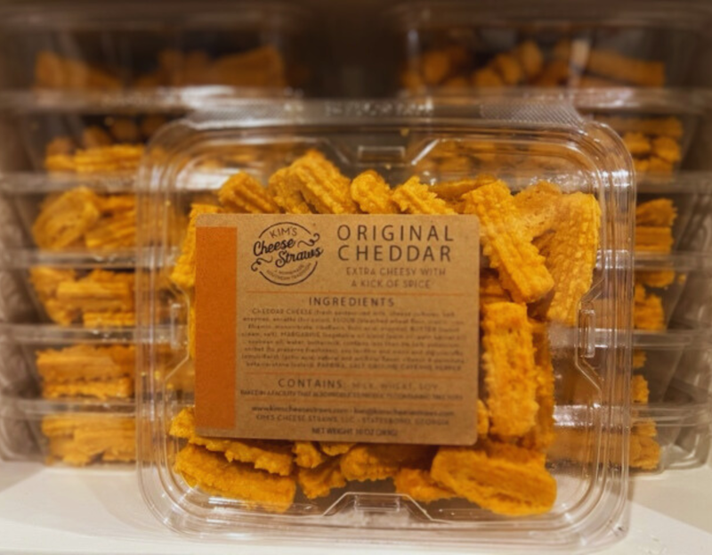 Close-up of a clear plastic container labeled “Kim’s Cheese Straws – Original Cheddar.” The container is filled with ridged, golden cheese straws and is stacked among several identical packages. The label notes the product is “extra cheesy with a kick of spice” and lists the ingredients.