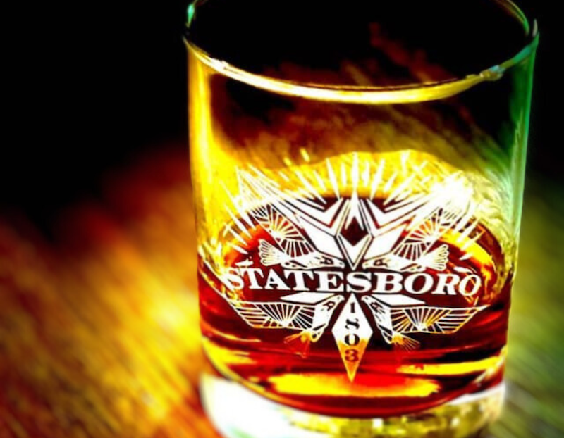 A close-up of a glass filled with a dark amber liquid, possibly whiskey, illuminated by warm lighting. The glass features an intricate white design with the word "STATESBORO" and the year "1803" prominently displayed in the center. The background is dark with a wooden surface faintly visible.