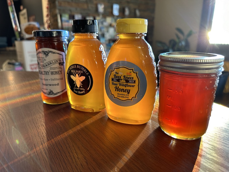 A variety of local honey jars and bottles displayed on a wooden counter. The collection includes four containers with labels from different producers, including Mr. Franklin's Healthy Honey, Humble Bumble Bee Farm, The Bee's Knees, and a mason jar of raw honey. Sunlight illuminates the golden hues of the honey, emphasizing their rich color and clarity.