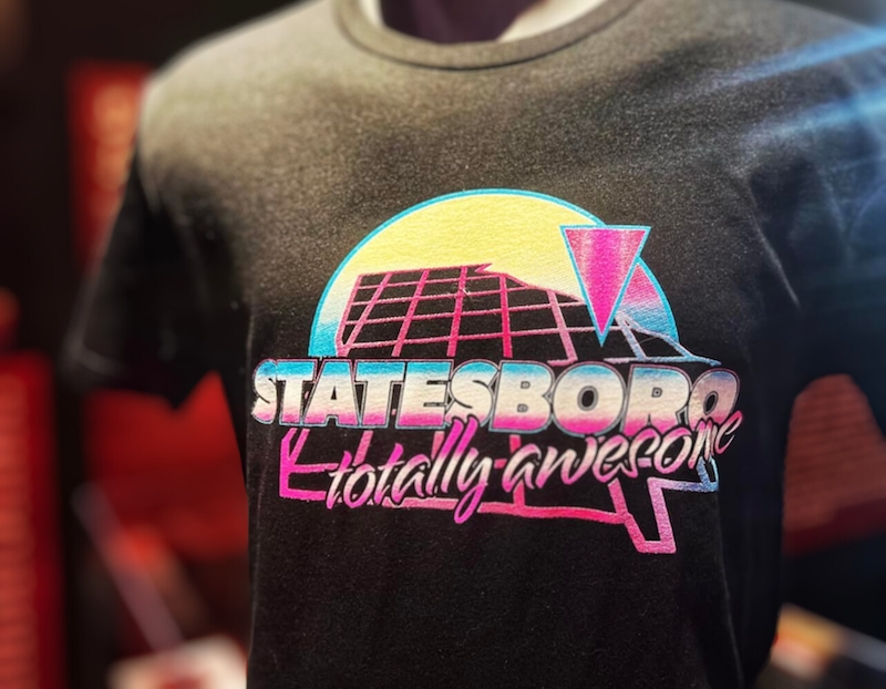 A black t-shirt featuring a vibrant retro design with the words "STATESBORO totally awesome" in bold, colorful font. The graphic includes a stylized grid, a yellow sun, and a pink triangle, reminiscent of 1980s aesthetics. The shirt is displayed on a mannequin inside the Visit Statesboro location.